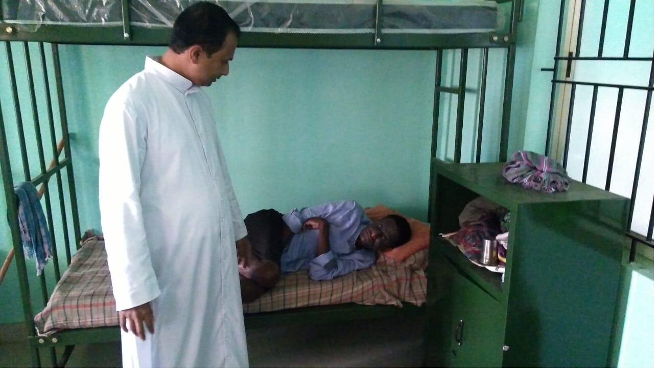 Father Jinto stands quietly beside a resident resting in his bed at the Karuna centre.