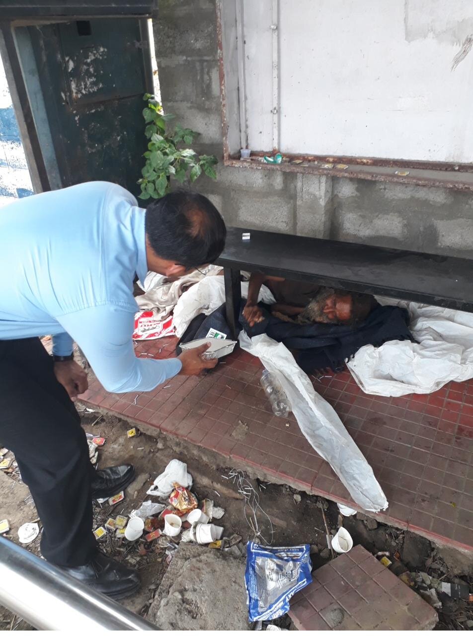 A Karuna volunteer kneels beside an elderly man resting under a bench in the street, offering him a hot meal.