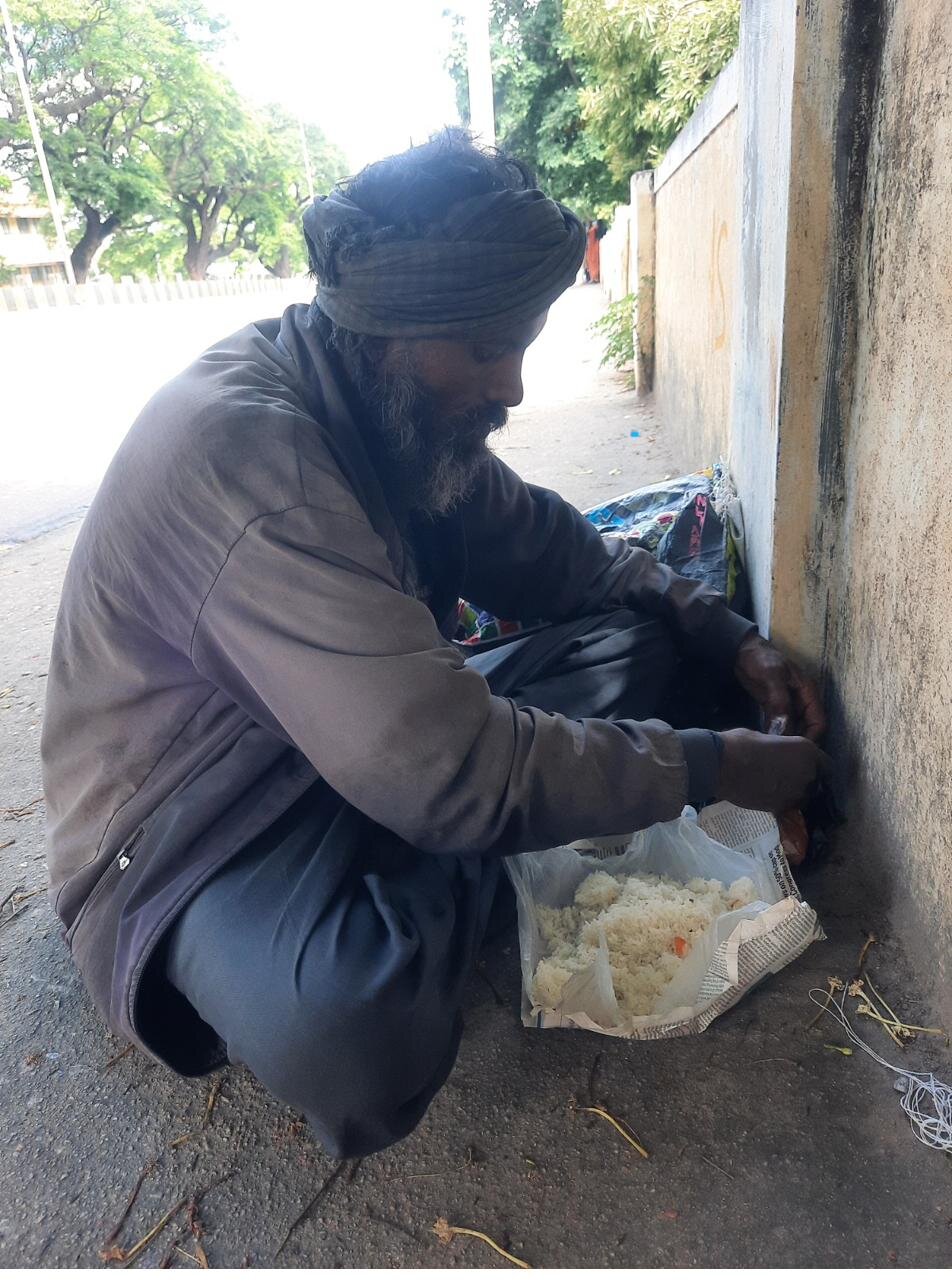 An elderly man eats rice from a newspaper plate, seated on a footpath.