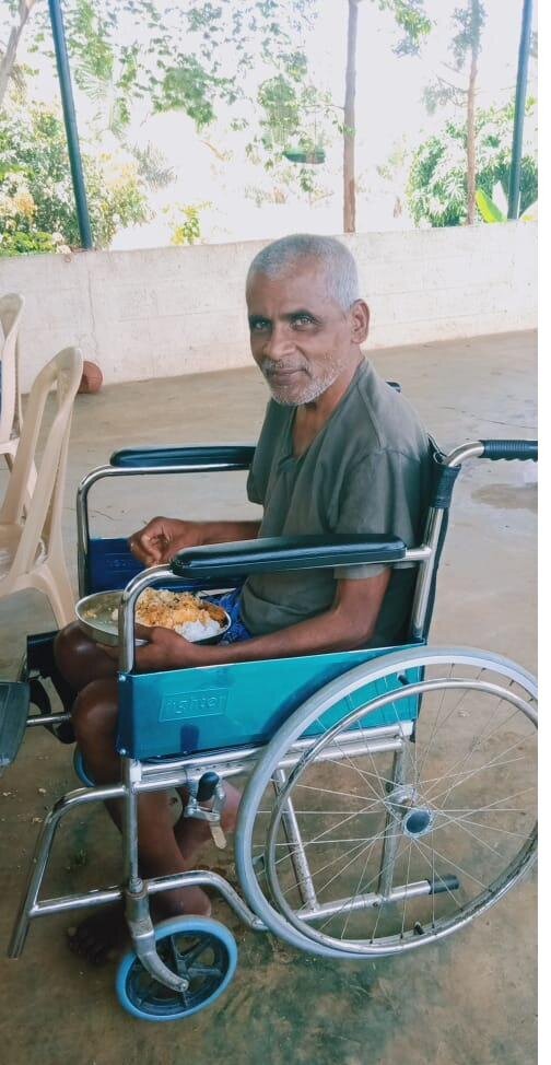 An elderly man sits in a wheelchair at the Karuna centre, smiling gently with a hot meal before him.